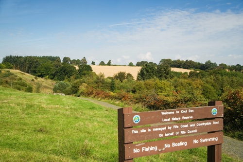 Photo of a green, grassy field with a sign stating "Welcome to Coul Den", trees in the distance and a clear blue sky.